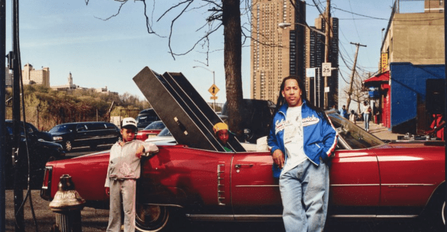 DJ Kool Herc standing in front of a open top car with two kids, with two big stereo speakers in the back seat 